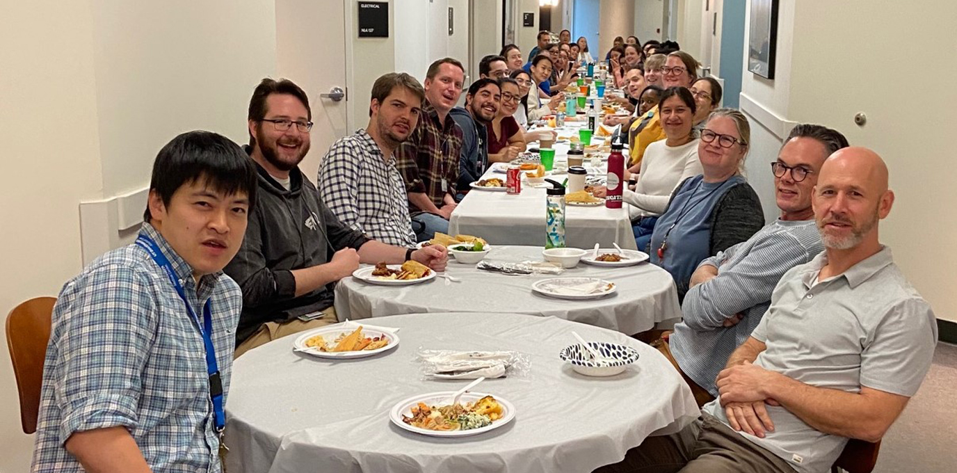 Group shot of approximately 35 people sitting at a long table with food and smiling for the camera