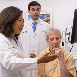 A woman and man with dark hair wearing white lab coats, work with a seated male patient with gray hair, wearing a peach shirt.