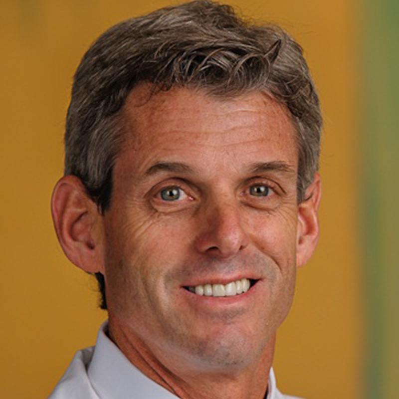man with brown-gray hair wearing a white dress shirt, yellow and blue striped tie, and white lab coat.