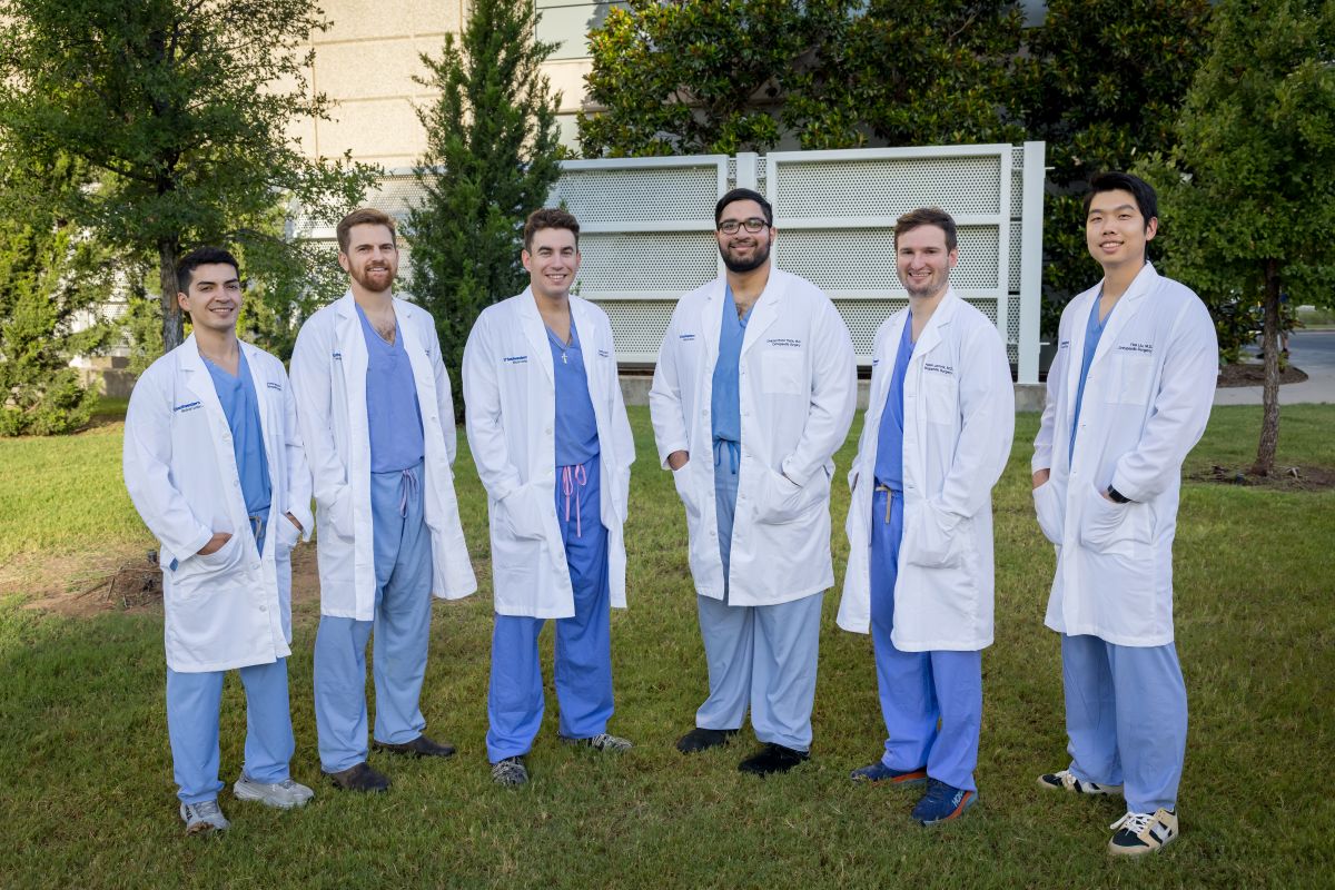 group of 6 male Orthopaedic Surgery Residents weariing white coats and blue scrubs standing outside hospital