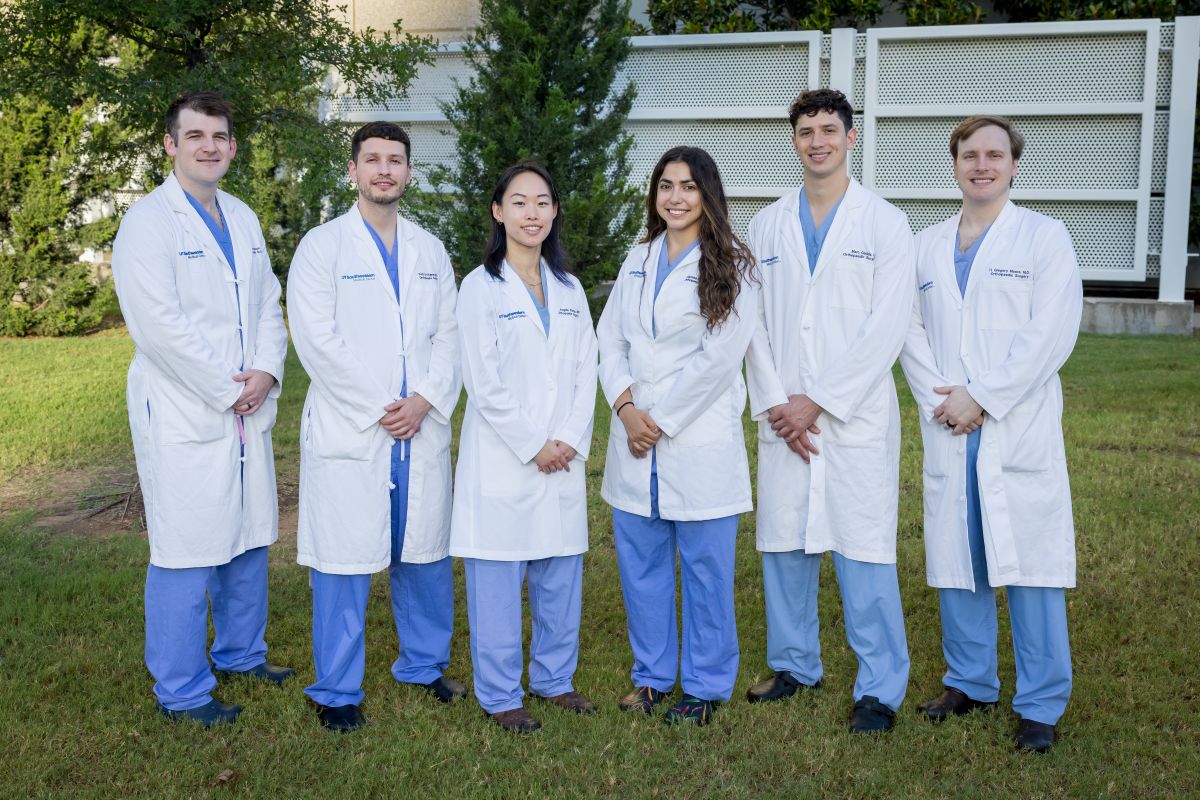 group of 4 men and 2 women Orthopaedic Surgery Residents weariing white coats and blue scrubs standing outside hospital