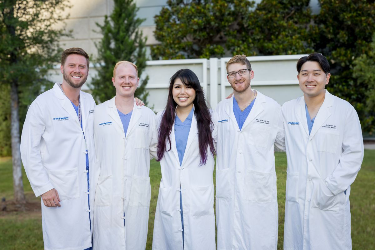 group of 4 men and 1 woman Orthopaedic Surgery Residents weariing white coats and blue scrubs standing outside hospital