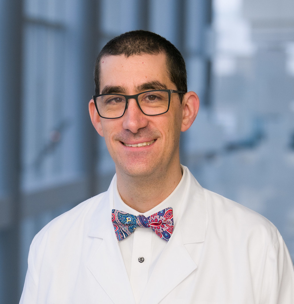 Dr. Sher in white coat, white shirt, and bowtie standing with windows in the background.
