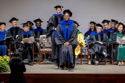 faculty and speakers in caps and gowns seated on stage behind standing man in glasses placing sash on shoulders of smiling black woman