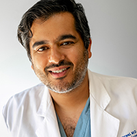 man with dark hair and grey beard wearing blue scrubs and a white coat