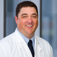 man with short brown hair wearing a light blue shirt, black tie and white coat with UT Southwestern Medical Center logo