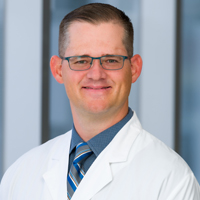 man with short brown hair wearing wire-rimmed glasses, a dark blue shirt, striped tie, and white coat