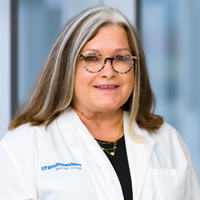 woman with shoulder-length brown hair with blonde streaks, wearing tortoise-shell glasses, a black shirt, and white coat