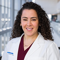 woman with long, dark, curly hair wearing a maroon shirt and white coat