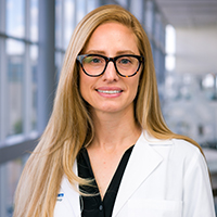 woman with long, blonde hair wearing tortoise-shell glasses, a black shirt, and white coat
