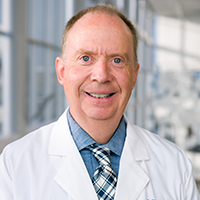 man with short, sandy blonde hair wearing a blue shirt, blue and white plaid tie, and white coat
