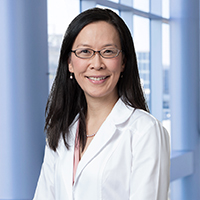 woman with long dark hair wearing a pink shirt, white coat, and black glasses