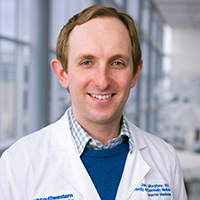 man with short, sandy hair wearing a blue plaid shirt, royal blue sweater, and white coat with the UT Southwestern Medical Center logo