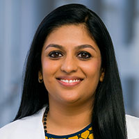 woman with long, dark hair wearing a gold and black patterned shirt and white coat