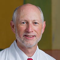 man with white hair and short, white beard wearing a white shirt, a red tie, and white coat