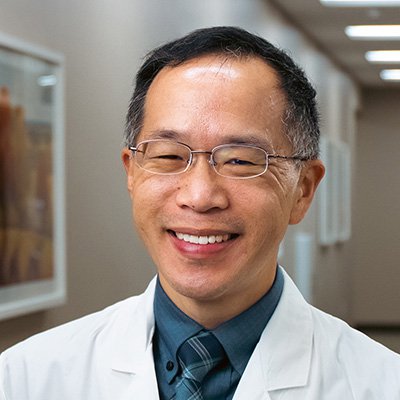 man with short dark hair wearing a dark teal shirt, teal and blue plaid tie, white coat, and wire-rimmed glasses