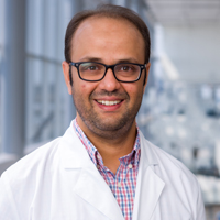 man with short brown hair wearing a red and blue checked shirt, white coat, and black glasses