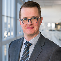 man with short brown hair wearing a light grey shirt, navy blue striped tie, dark grey coat, and tortoiseshell glasses