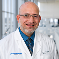 bald man with short grey beard wearing a royal blue shirt, blue and orange striped tie, and wire-rimmed glasses