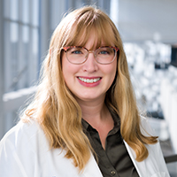 woman with long strawberry blonde hair and bangs wearing a dark olive green shirt, white coat, and wire-rimmed glasses