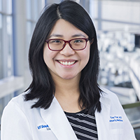 woman with shoulder-length black hair wearing a black and white striped shirt, white coat, and red glasses