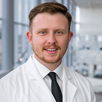 man with red hair and a short beard wearing a white shirt, black tie, and white coat
