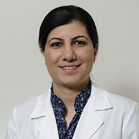 woman with straight black hair pulled back in a bun wearing a dark blue patterned shirt and white coat