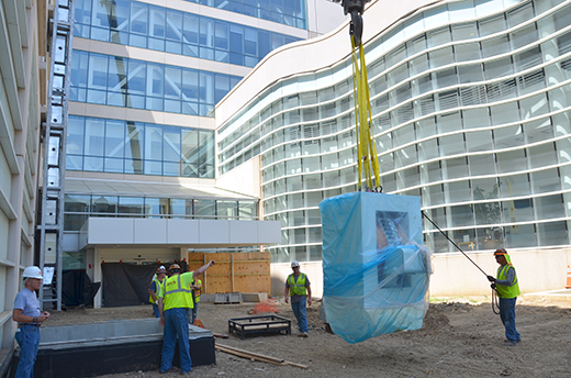 Installation of Cyclotron Construction workers guide heavy machine being carried by crane