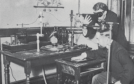 Taking an X-ray image with early Crookes tube apparatus Historic photo of one man holding his hand on an X-ray plate while another operates machinery