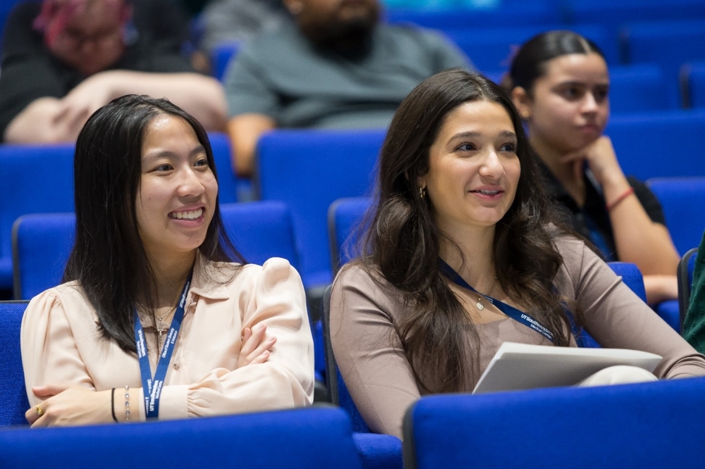 Research & Health Careers Conference for students interested in a career in medicine and science. 3 female undergraduate students sit in the audience (foreground) - from left, young Asian woman in pink shirt, woman with long dark hair in brown shirt, and in behind her woman with dark hair in a sleek bun wears a black shirt