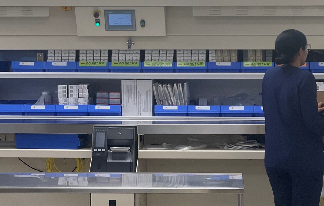 moody black and white image of person facing away in front of 2 walls of shelves full of medical drug boxes with empty metal exam table in the foreground