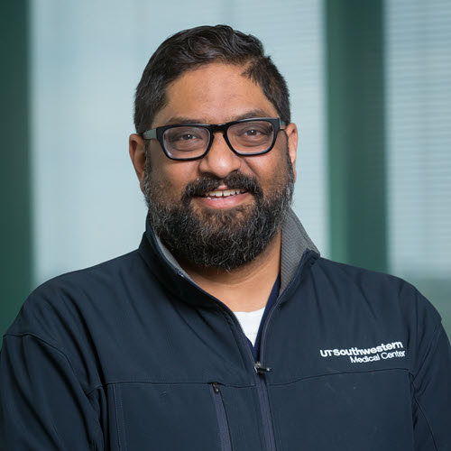 Smiling man with dark curly hair and a dark beard and mustache, wearing a blue polo shirt with the UT Southswestern Medical Center logo.