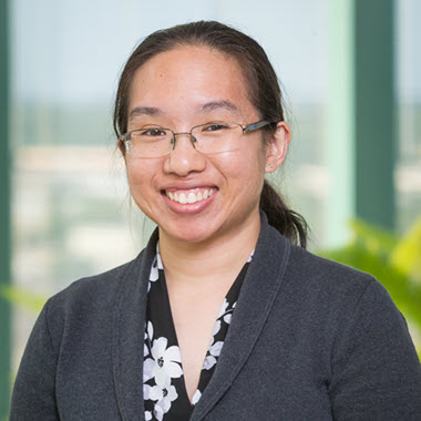 Smiling woman with long black hair pulled back in a pony tail, wearing a dark jacket, print blouse, and light-framed glasses.