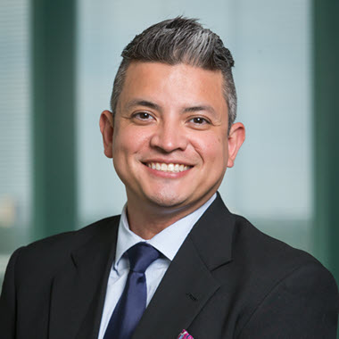 Smiling man with short graying hair, wearing a dark suit over a light dress shirt and blue tie.
