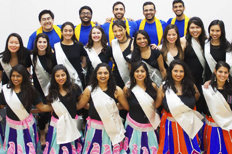 A group of dancers. Women dancers wear black tops with pink and blue skirts and white sashes and men wear blue shirts with yellow sashes.