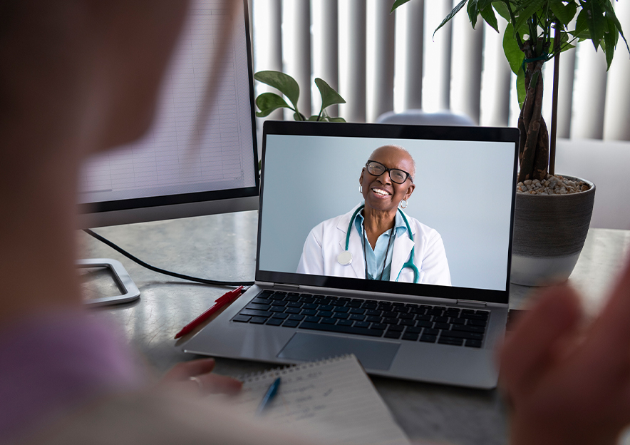 A Black doctor in a white coat with a stethoscope around his neck appears on a computer monitor