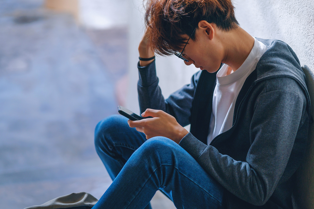 An upset teenage boy student, dressed in casual outfit, sits on a stairway reading on his smartphone with a backpack beside him, while dealing with the issue of cyberbullying.