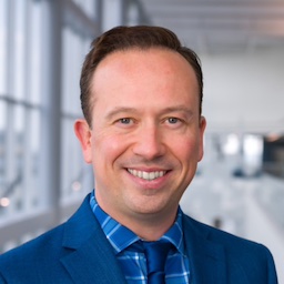 Dr. Lutz headshot: white man smiling, wears blue blazer, plaid blue shirt and blue tie, atrium background