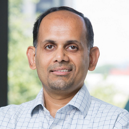 Dr. Binu Thomas wearing a plaid blue and white shirt standing in front of a window