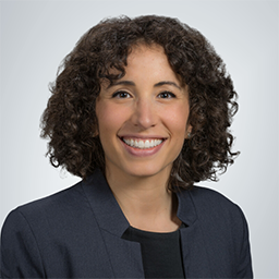 Dr. Ana Valencia wearing a dark grey jacket with a black shirt on a white background